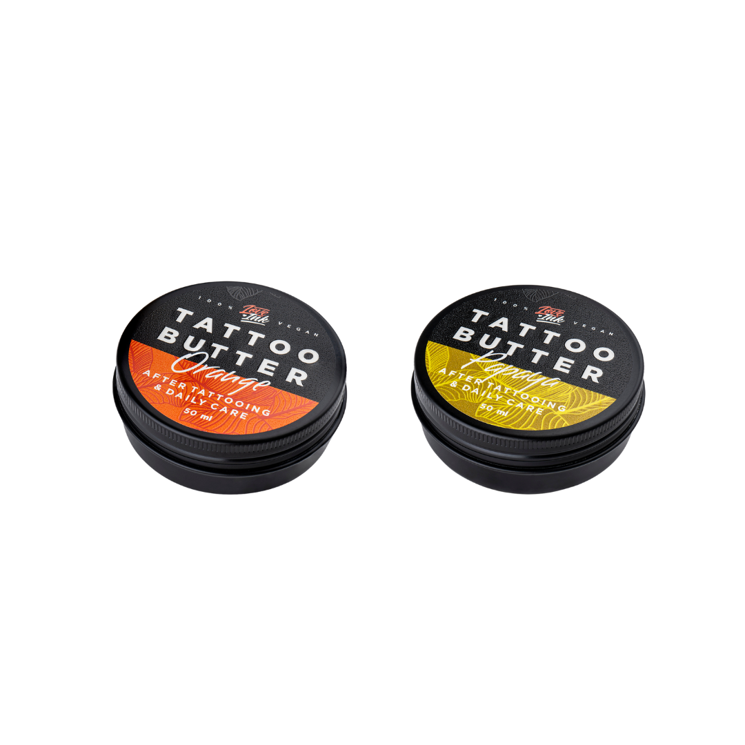 Two tins of Love Ink Tattoo Butter on a transparent background. One tin is labeled 'Tattoo Butter Orange' with an orange design, and the other is labeled 'Tattoo Butter Papaya' with a yellow design. Both tins are 50 ml and are intended for after tattooing and daily care.