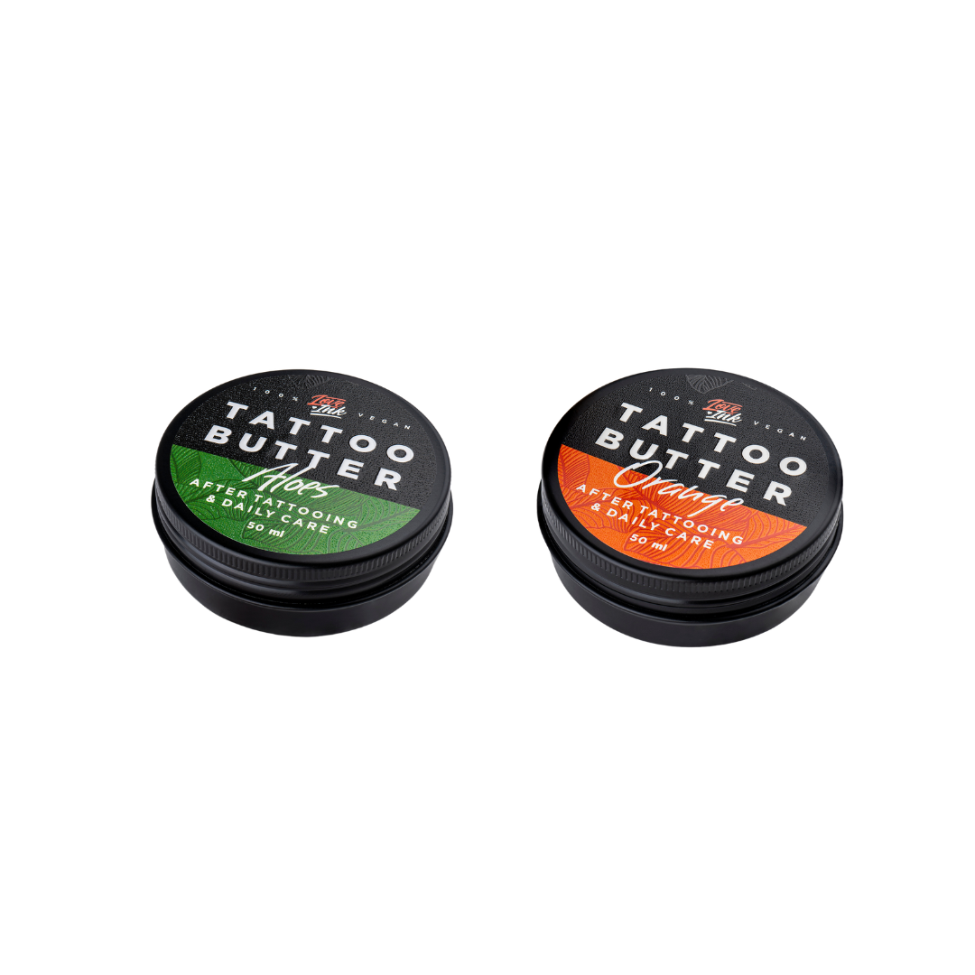 Two tins of Love Ink Tattoo Butter on a transparent background. One tin is labeled 'Tattoo Butter Aloes' with an green design, and the other is labeled 'Tattoo Butter Orange' with an orange design. Both tins are 50 ml and are intended for after tattooing and daily care.