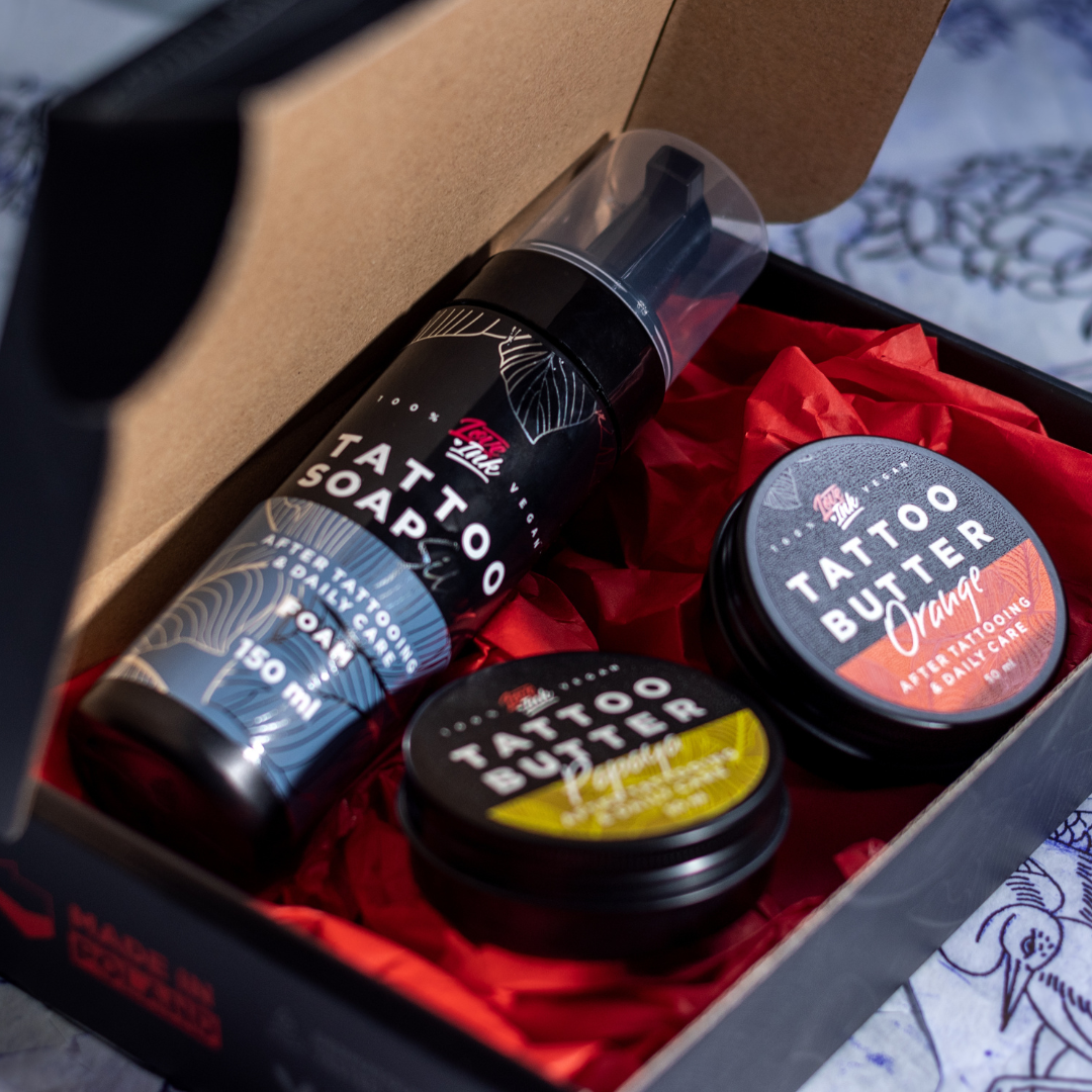 An open box containing Love Ink tattoo care products, including a bottle of Tattoo Soap and two tins of Tattoo Butter (Papaya and Orange), placed on red tissue paper. The background features various tattoo sketches and designs