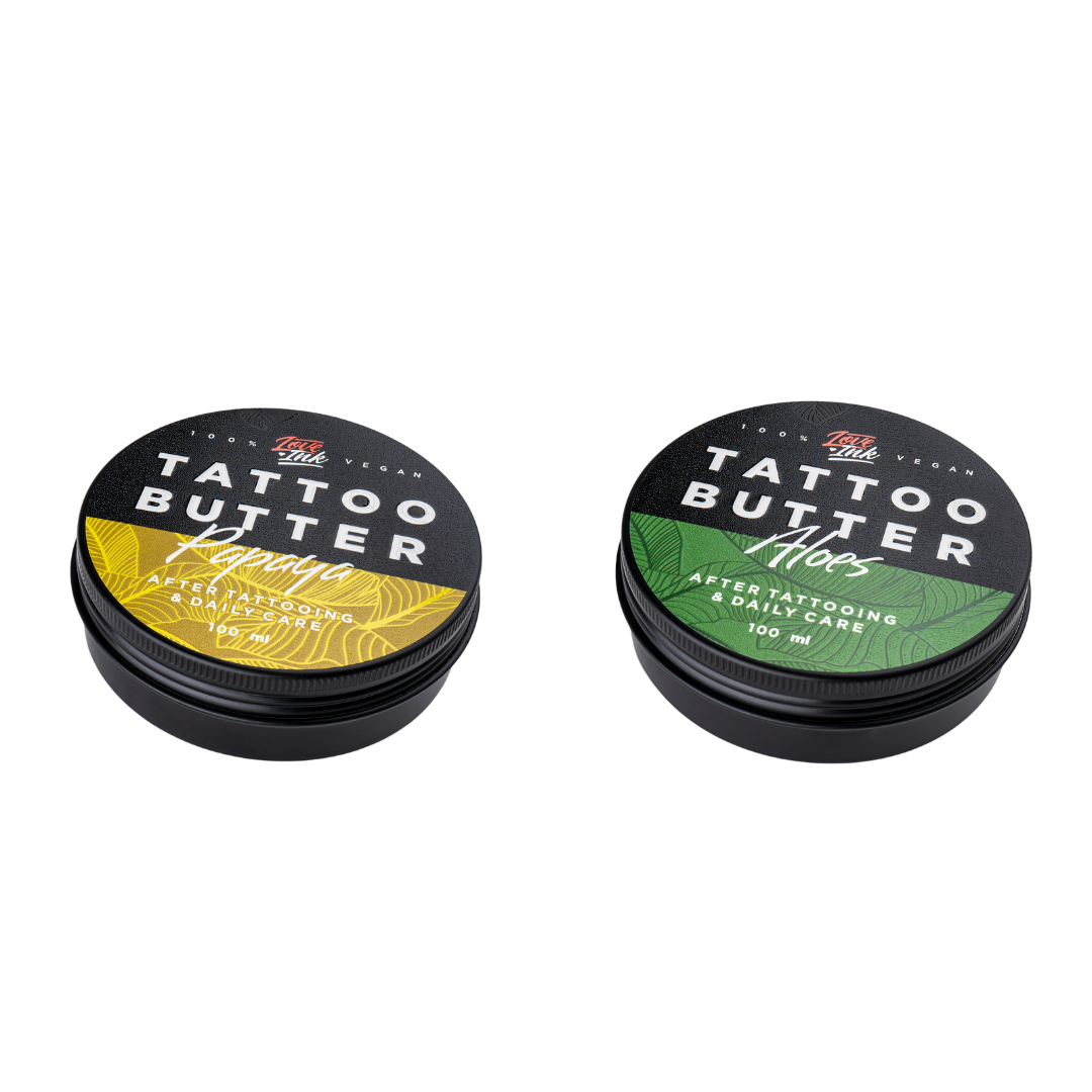 Two tins of Love Ink Tattoo Butter on a transparent background. One tin is labeled 'Aloe' with a green design, and the other is labeled 'Papaya' with an yellow design. Both tins are 100 ml and are intended for after tattooing and daily care.