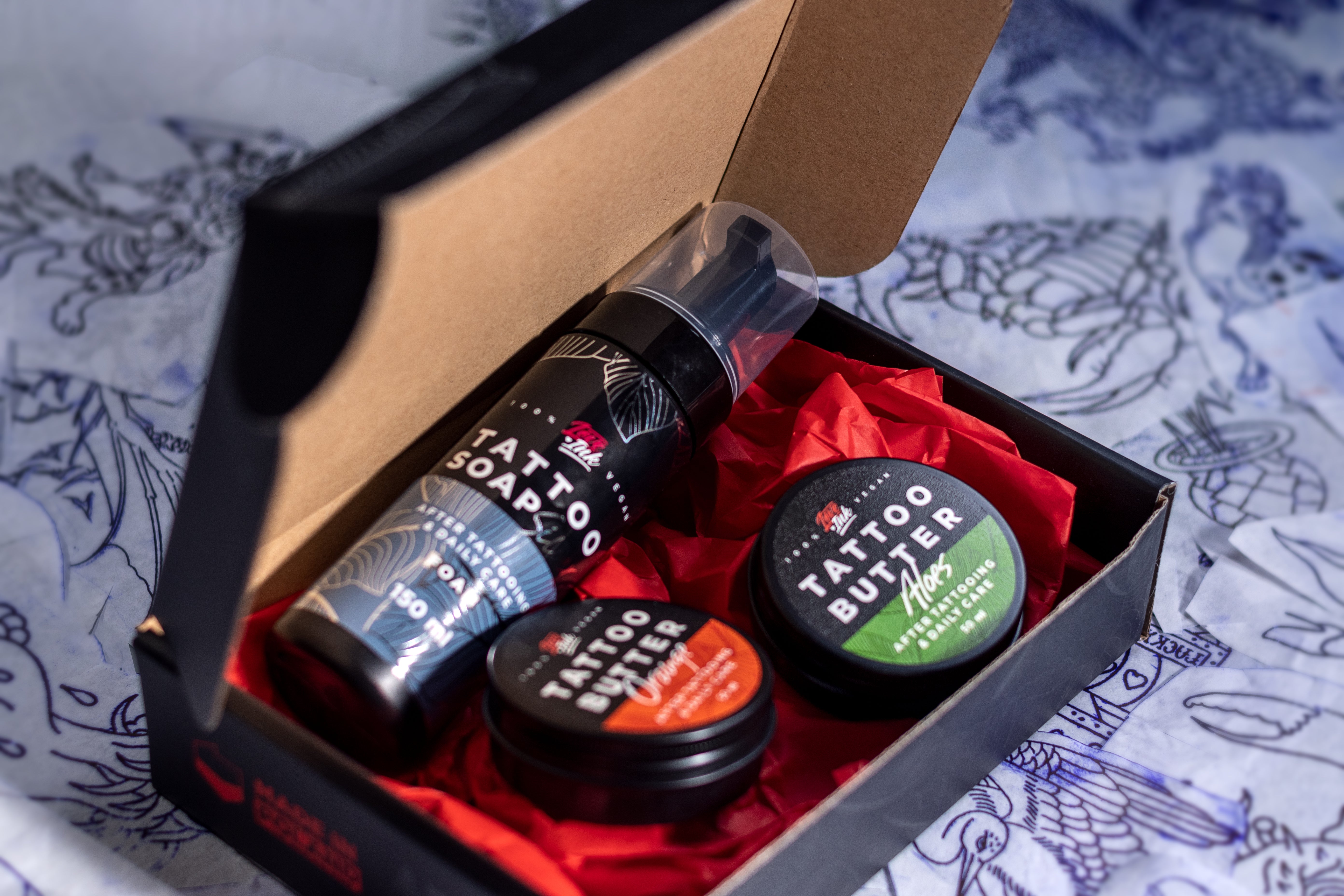 An open box containing Love Ink tattoo care products, including a bottle of Tattoo Soap and two tins of Tattoo Butter (Orange and Aloe), placed on red tissue paper. The background features various tattoo sketches and designs