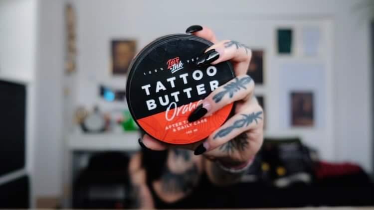 Hand holding a jar of Tattoo Butter Orange by LoveInk, designed for after tattooing and daily care. The background shows a cozy room with blurred details, emphasizing the product.