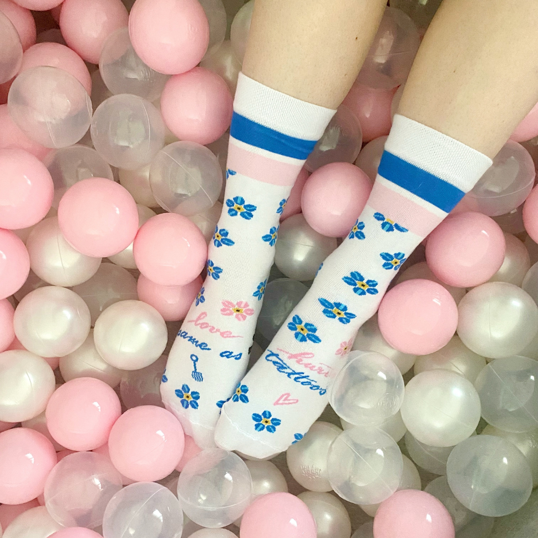 Feet wearing white socks with blue and yellow flower patterns, blue stripes at the top, and the Love Ink logo, lying in a ball pit filled with pink and white balls.