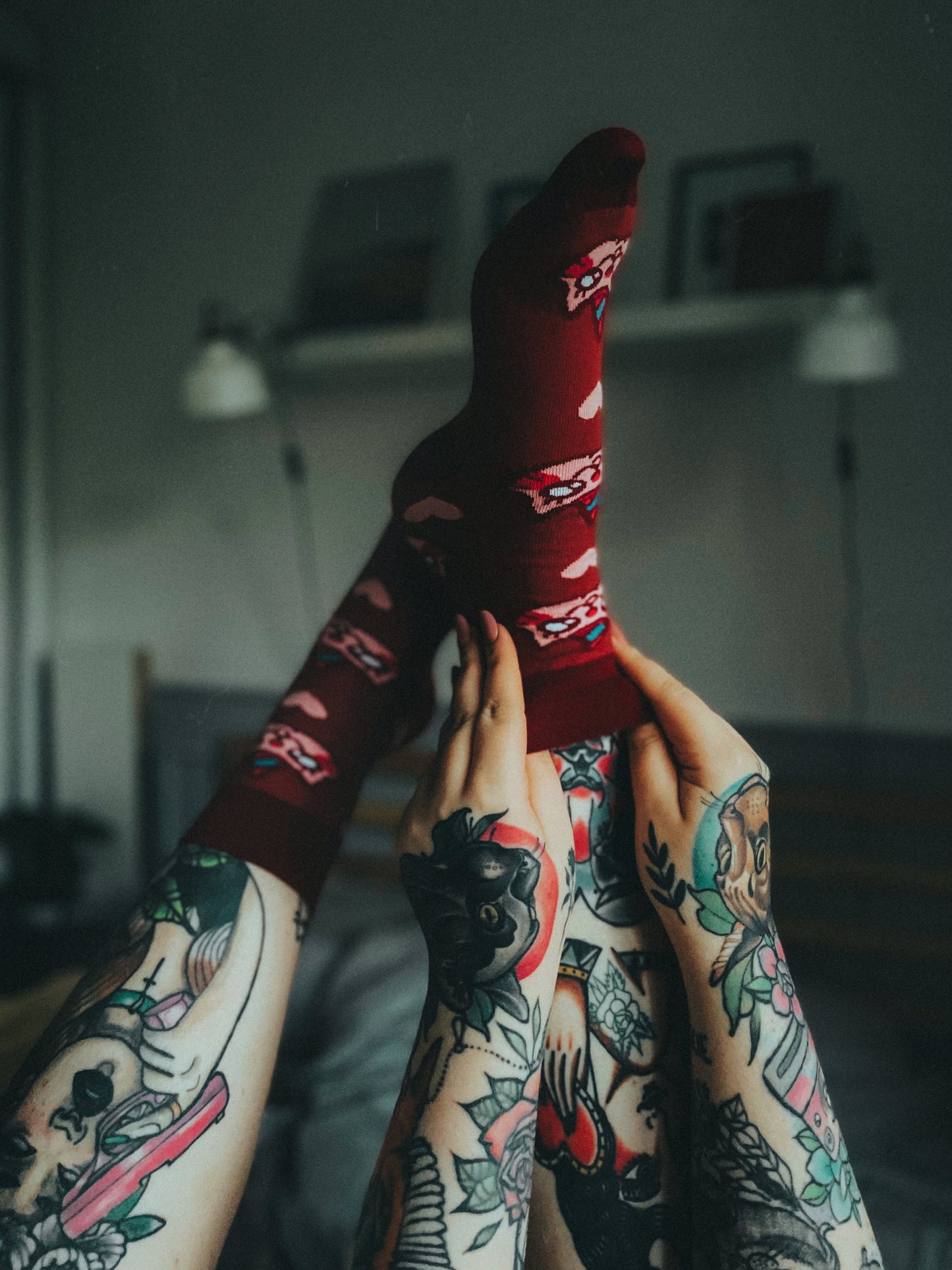 Tattooed legs raised with feet wearing red socks featuring a cartoon cat with a unicorn horn (Kotorozec) and hearts, set in a cozy indoor setting.