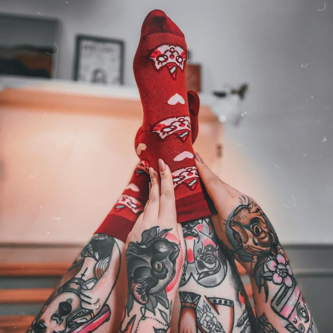Tattooed legs with feet wearing red socks featuring a cartoon cat with a unicorn horn (Kotorozec) and hearts, posed against a blurred indoor background.