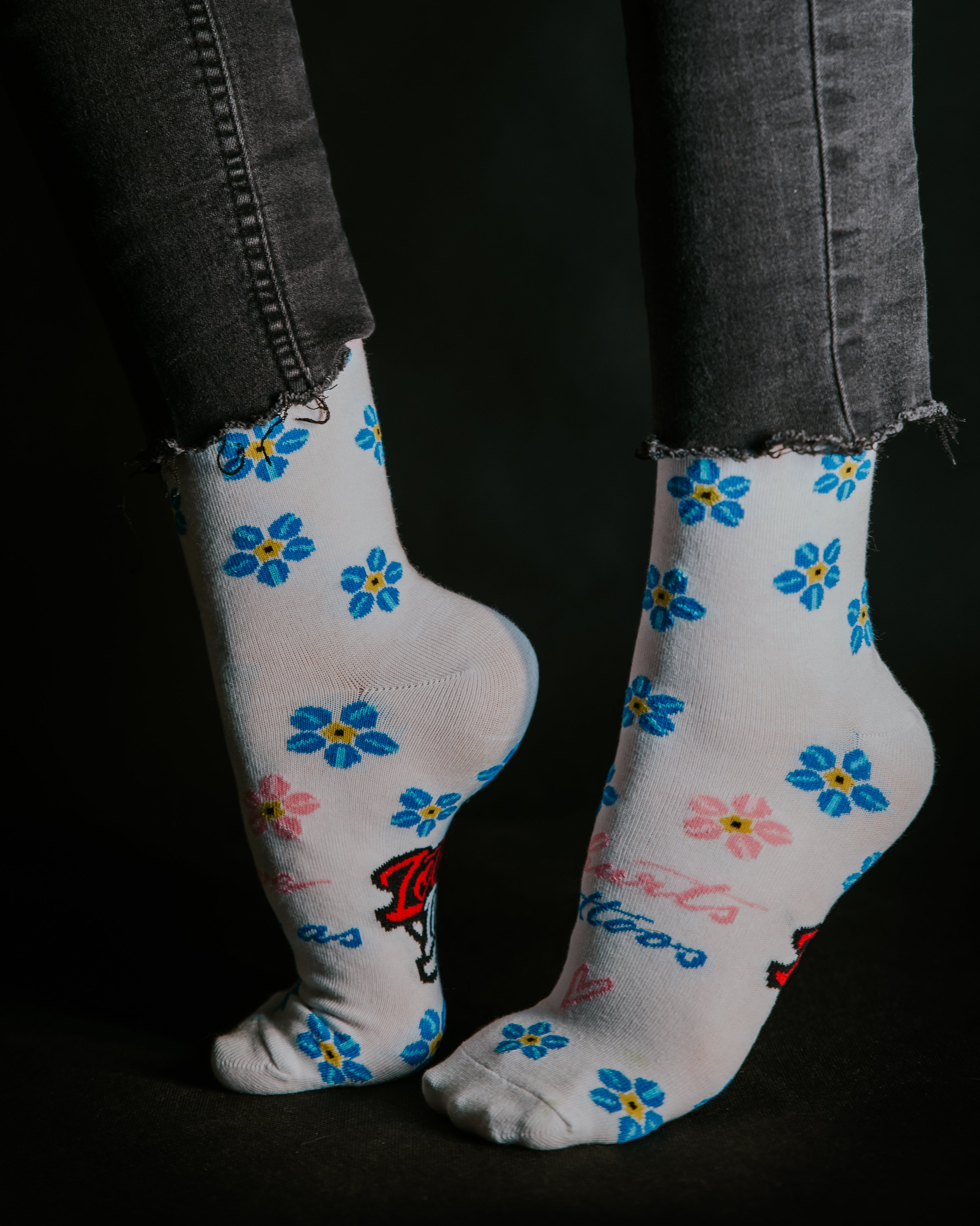 Close-up of feet wearing white socks with blue and yellow flower patterns and the Love Ink logo, paired with frayed-edge black jeans, set against a dark background.