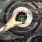 Hand holding a tin of Love Ink Tattoo Butter Aloe, 50 ml, in front of a black and white moon-themed tapestry.