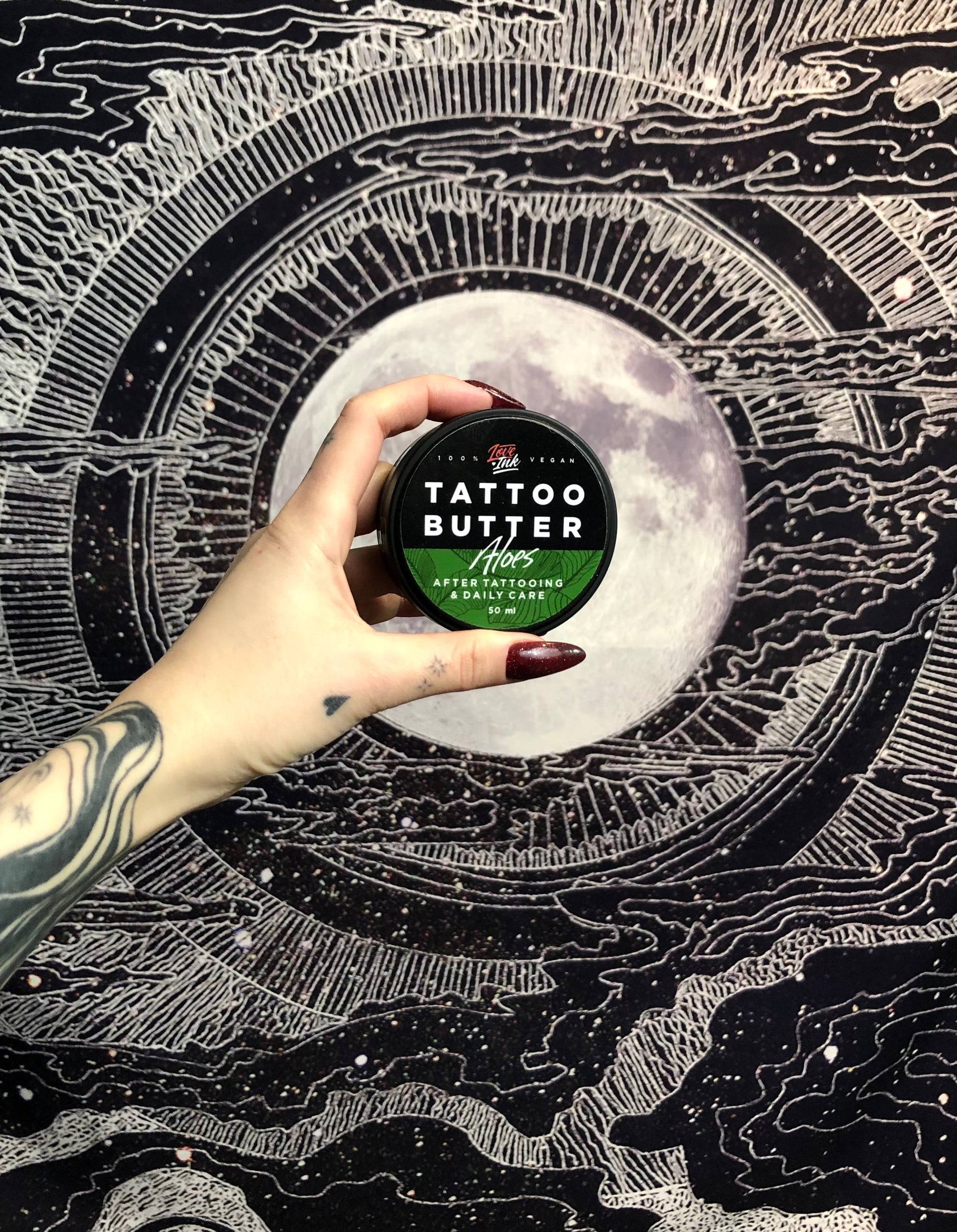 Hand holding a tin of Love Ink Tattoo Butter Aloe, 50 ml, in front of a black and white moon-themed tapestry.