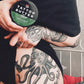 Tattooed person holding a tin of Love Ink Tattoo Butter Aloe, with detailed tattoos visible on their arm and leg.