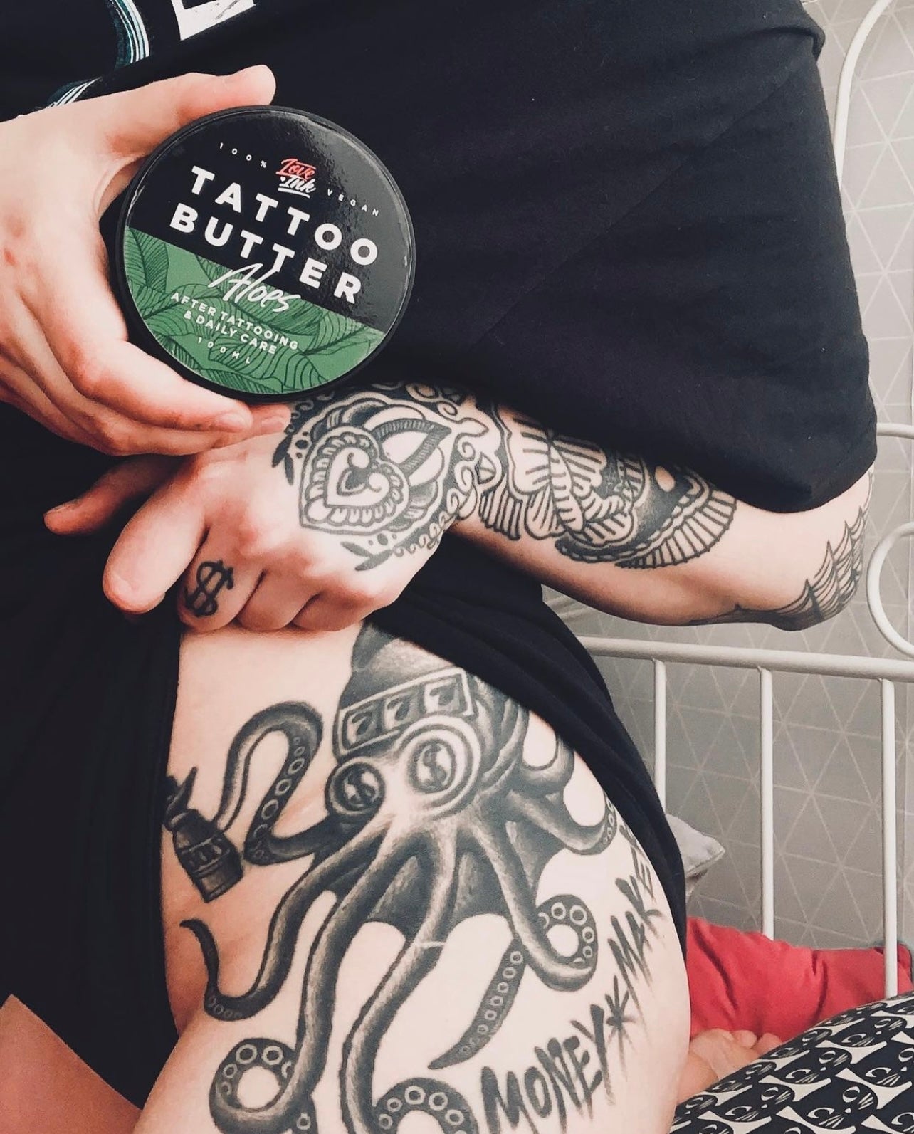 Tattooed person holding a tin of Love Ink Tattoo Butter Aloe, with detailed tattoos visible on their arm and leg.