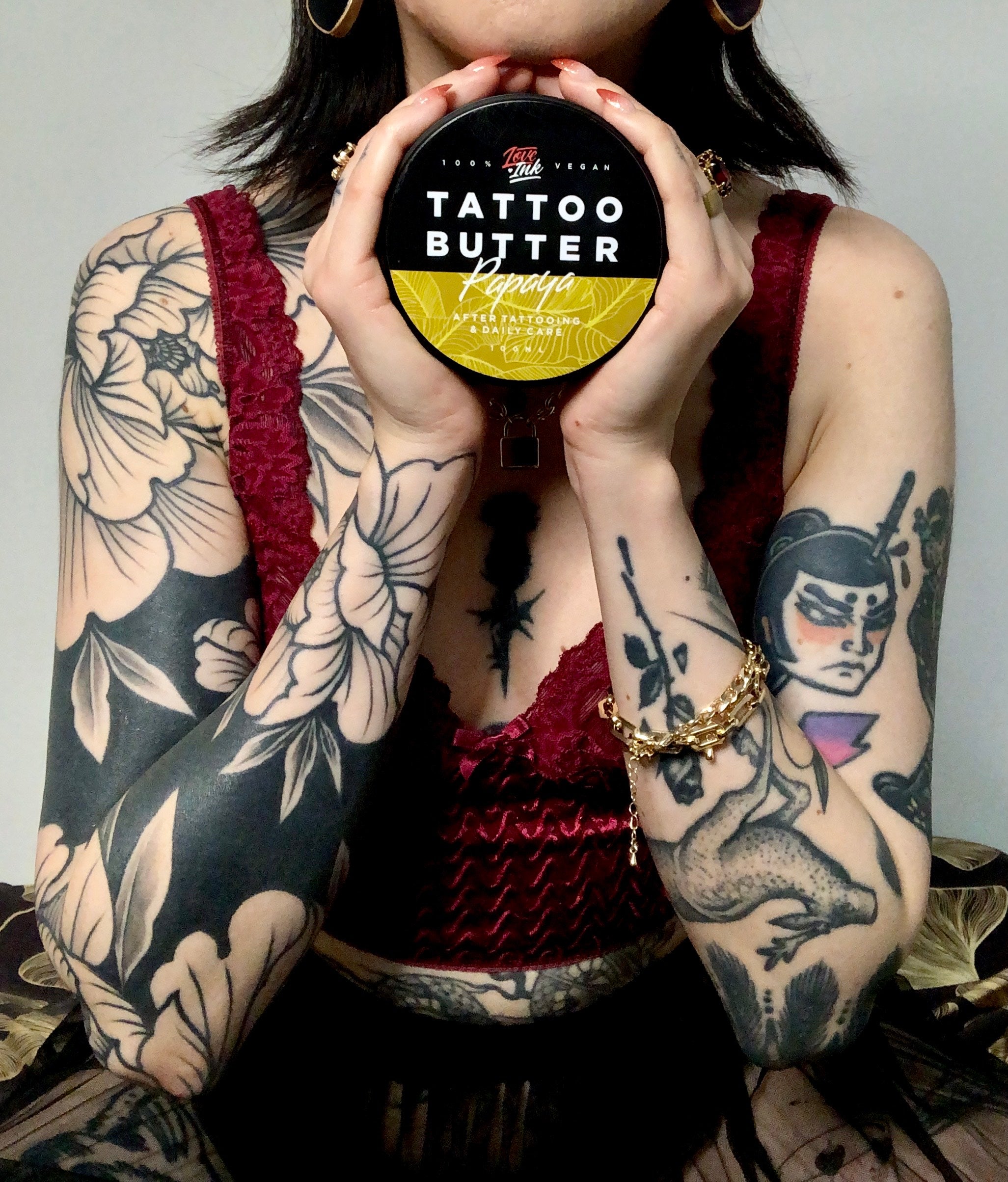 Person holding a jar of Tattoo Butter Papaya by LoveInk with detailed arm tattoos, showcasing the product's relevance for tattoo care.