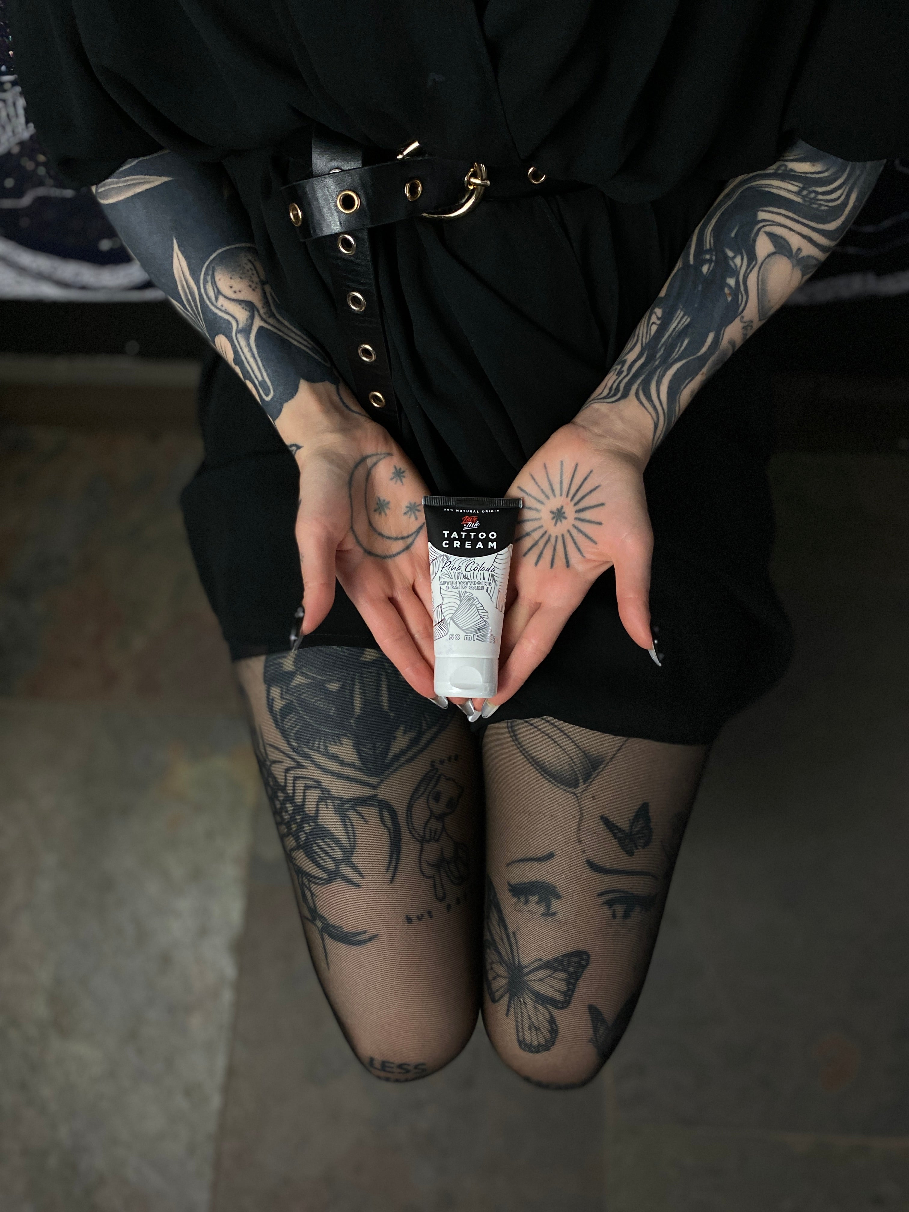 Person in a black outfit holding a tube of Tattoo Cream by LoveInk, showcasing detailed tattoos on arms and legs