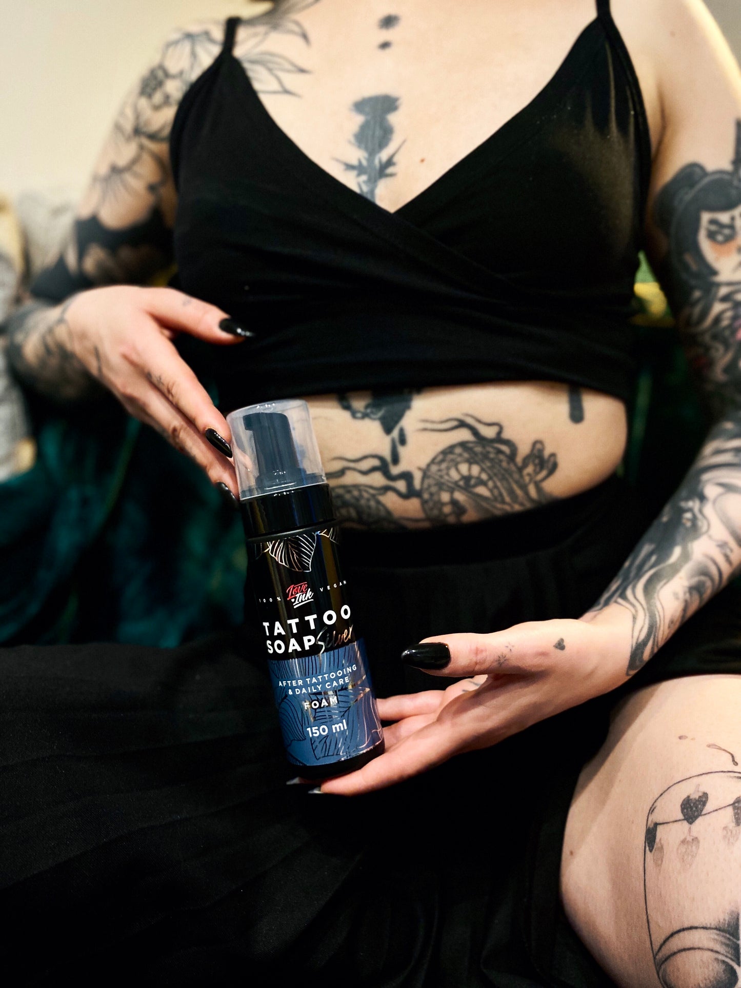 Woman with tattoos holding LoveInk's Tattoo Soap Silver bottle, showcasing tattoos on her arms, stomach, and legs. The product is for tattoo care and comes in a 150ml bottle.