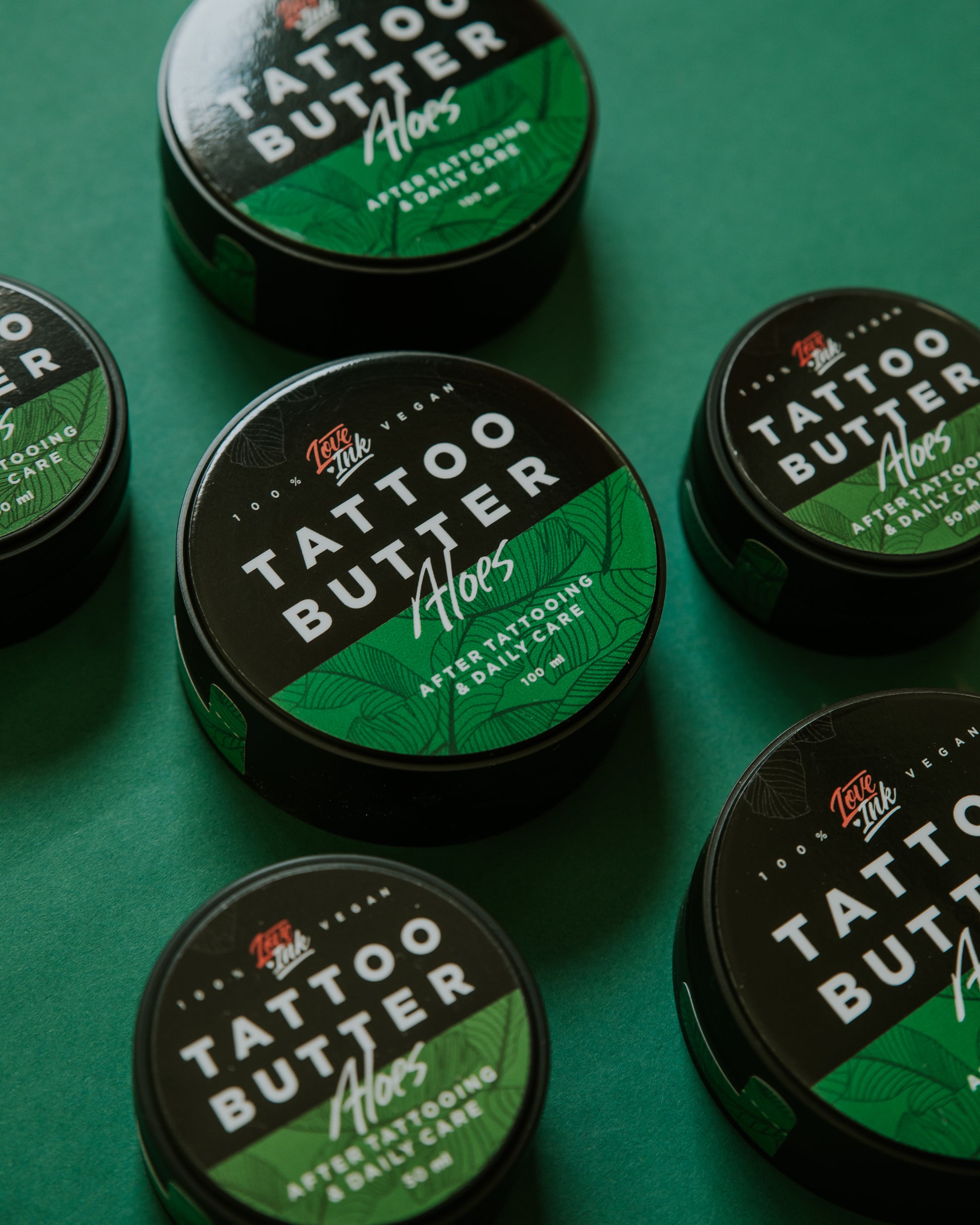 Multiple tins of Love Ink Tattoo Butter Aloe arranged on a green surface.