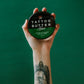 A hand holding a tin of Love Ink Tattoo Butter Aloe against a green background, with a floral tattoo visible on the wrist.